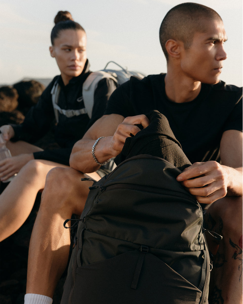 A man taking out a coat from the main compartment on his Motion Adventure Pack 38L in Charcoal with a woman sitting beside him with a backpack on her back