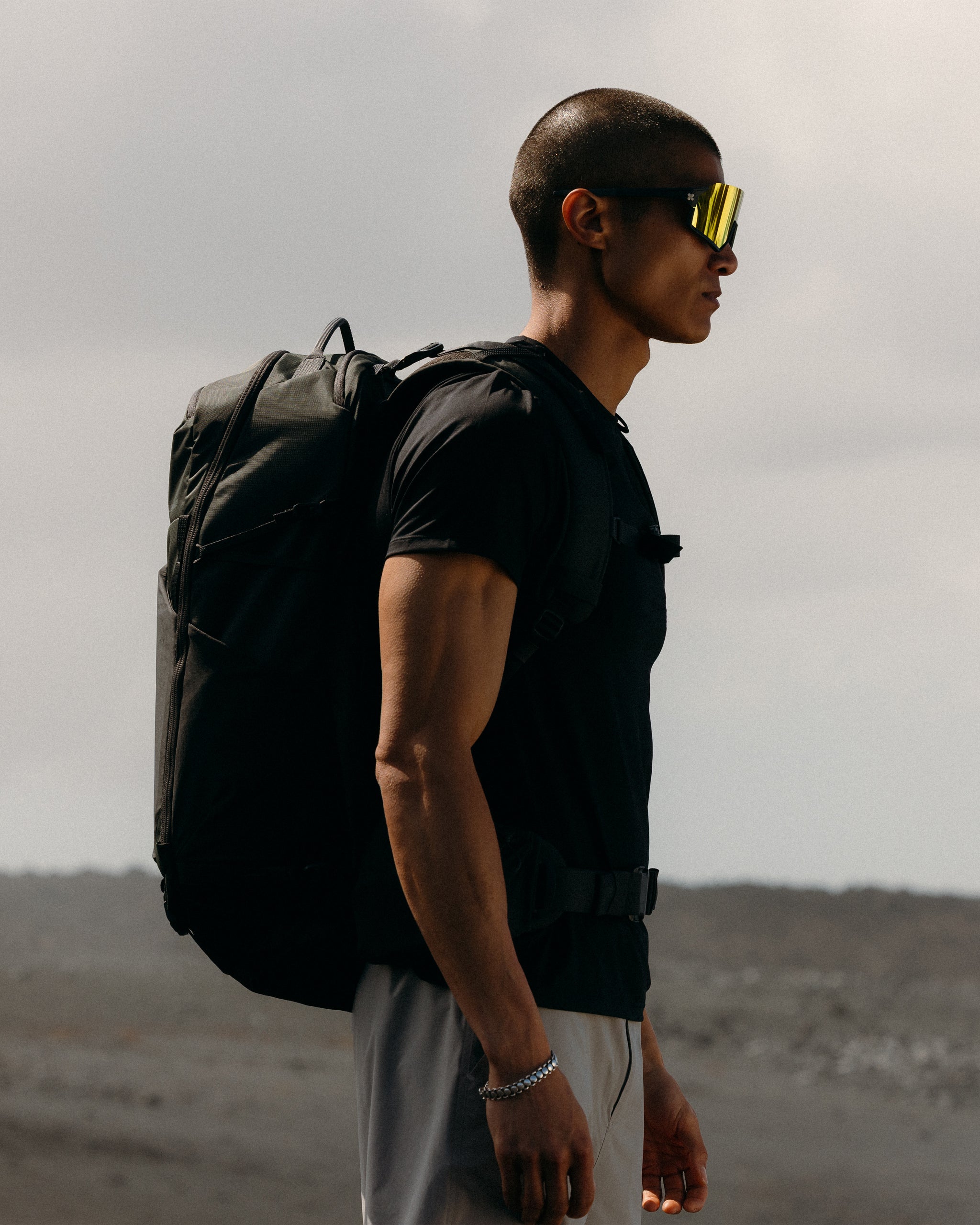 A man standing side on with sunglasses on and wearing a Motion Adventure Pack 38L in Charcoal on his back