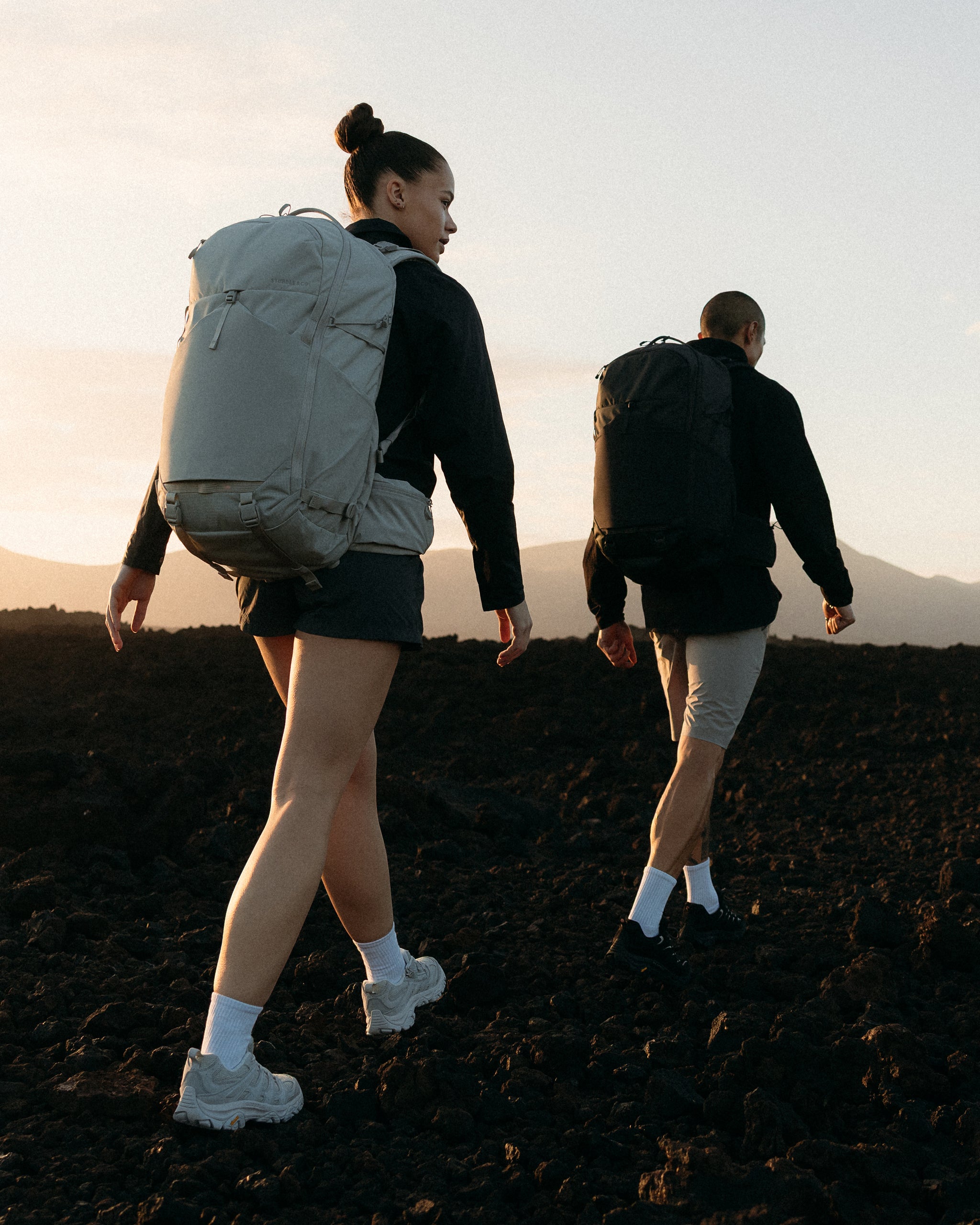 A man and woman walking across a rocky landscape at sunset with Motion Adventure Pack 38Ls on their backs in Ash and charcoal