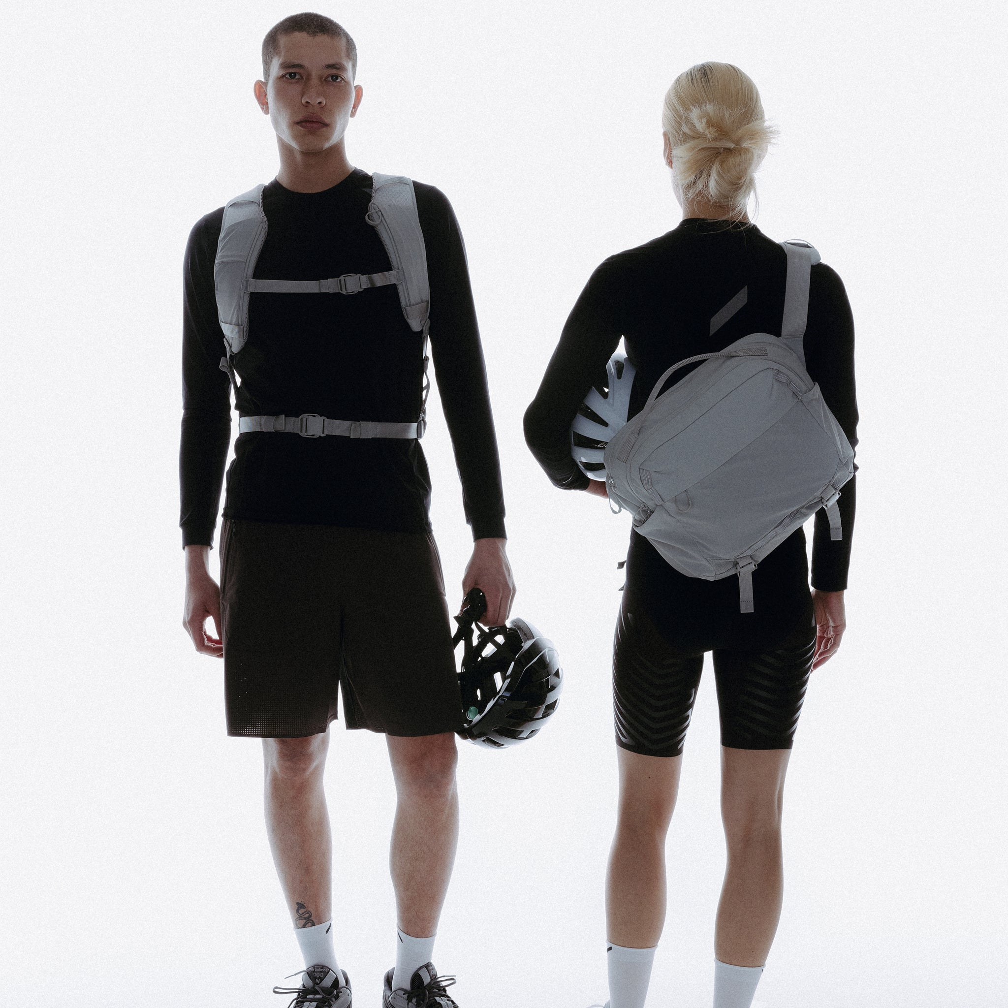 A man facing the camera and a woman facing a way, both holding bike helmets and wearing motion bags in Ash