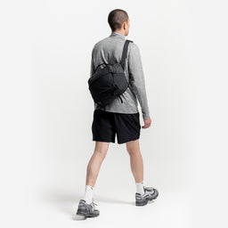 A studio shot of a man walking wearing a Charcoal Motion Sling 15L on his back