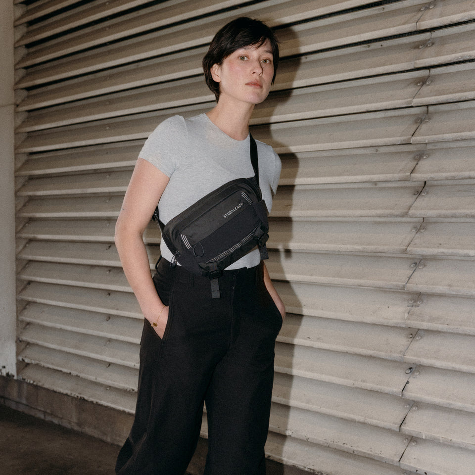 A woman standing in front of a metal garage with a charcoal motion sling 4l across her chest using the crossbody strap