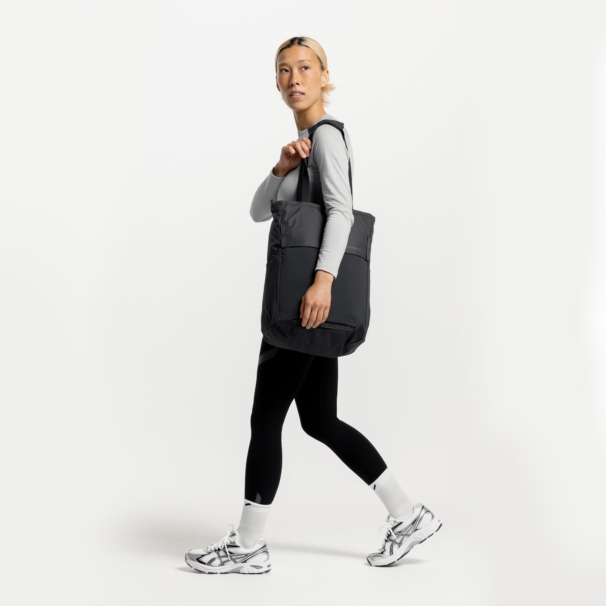 A studio shot of a woman walking with a Charcoal Motion Tote Pack 18L on her shoulder