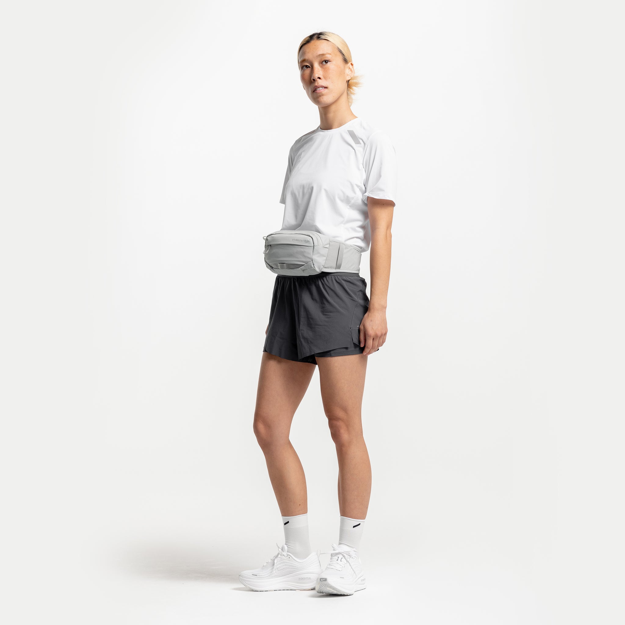 A studio shot of a woman wearing the Ash Motion Waist Pack 2L across her front on her waist