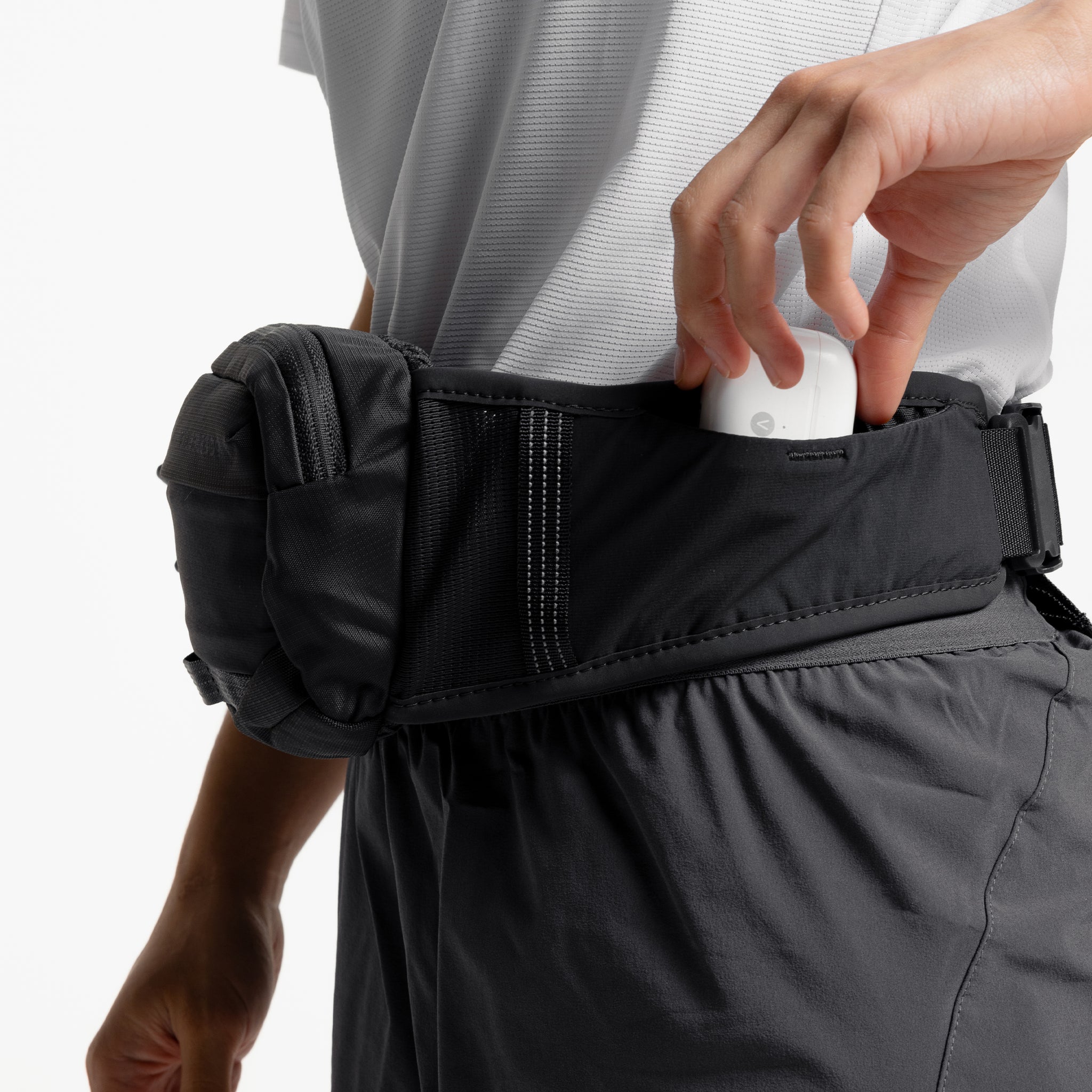 A close up shot of airpods being placed into the pocket on the waist strap of the Motion Waist Pack 2L in Charcoal, Ash and Magma