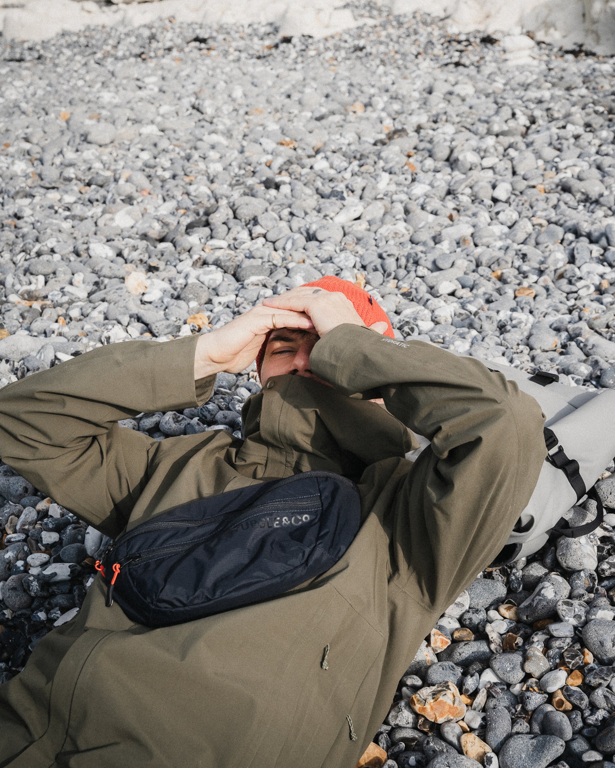a man lying on a pebble beach wearing a black packable sling- the travel project blog
