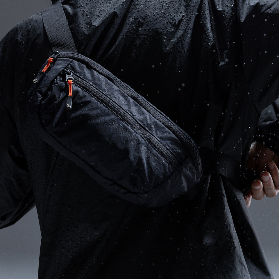 A black waterproof crossbody waist bag with orange zipper pulls, worn across the chest of a person wearing a black rain jacket.