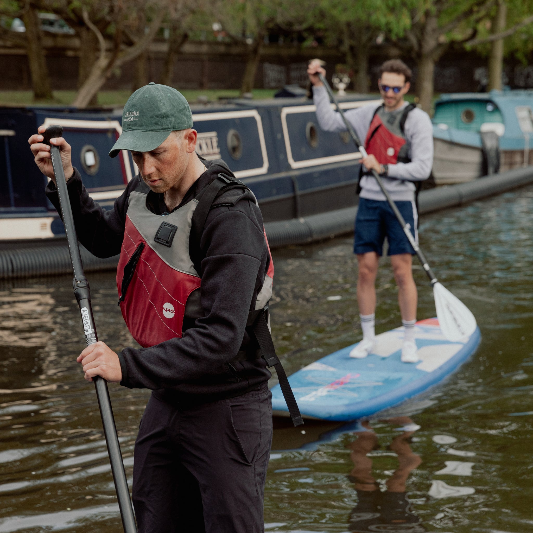 2 men paddle boarding up the river by canal boats