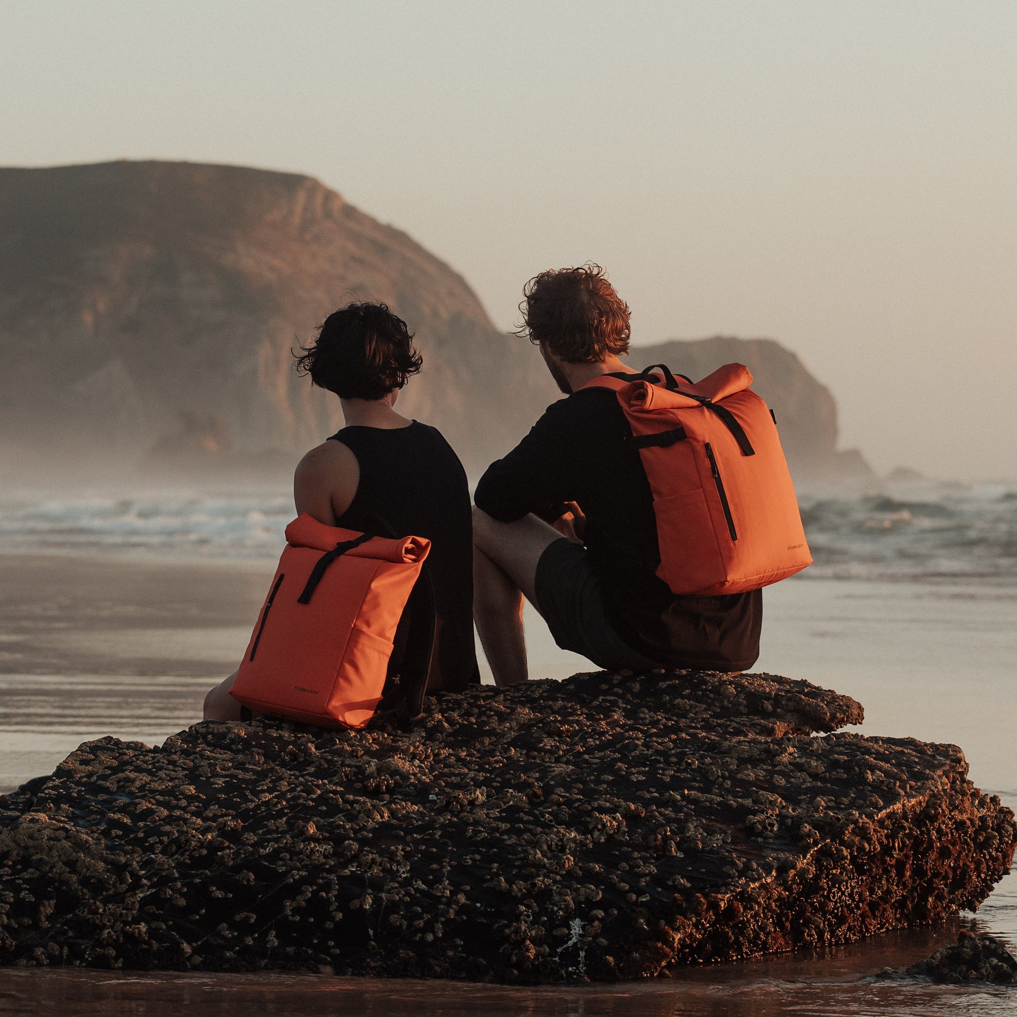 Man and women wearing The Roll Top 15L in Ember Orange on a beach
