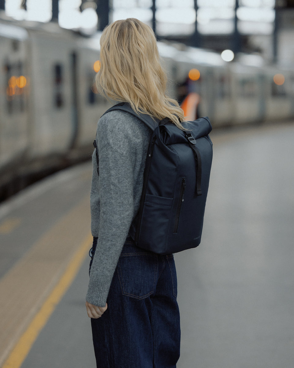 A woman wearing a Roll Top 15L on her back whilst standing on a train platform at the station