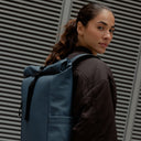 A woman standing side on wearing a Roll Top 15L backpack on her back in Tasmin Blue