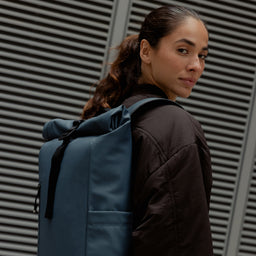 A woman standing side on wearing a Roll Top 15L backpack on her back in Tasmin Blue