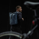 A man standing next to a bike with a Roll Top 15L backpack in Tasmin Blue on his back