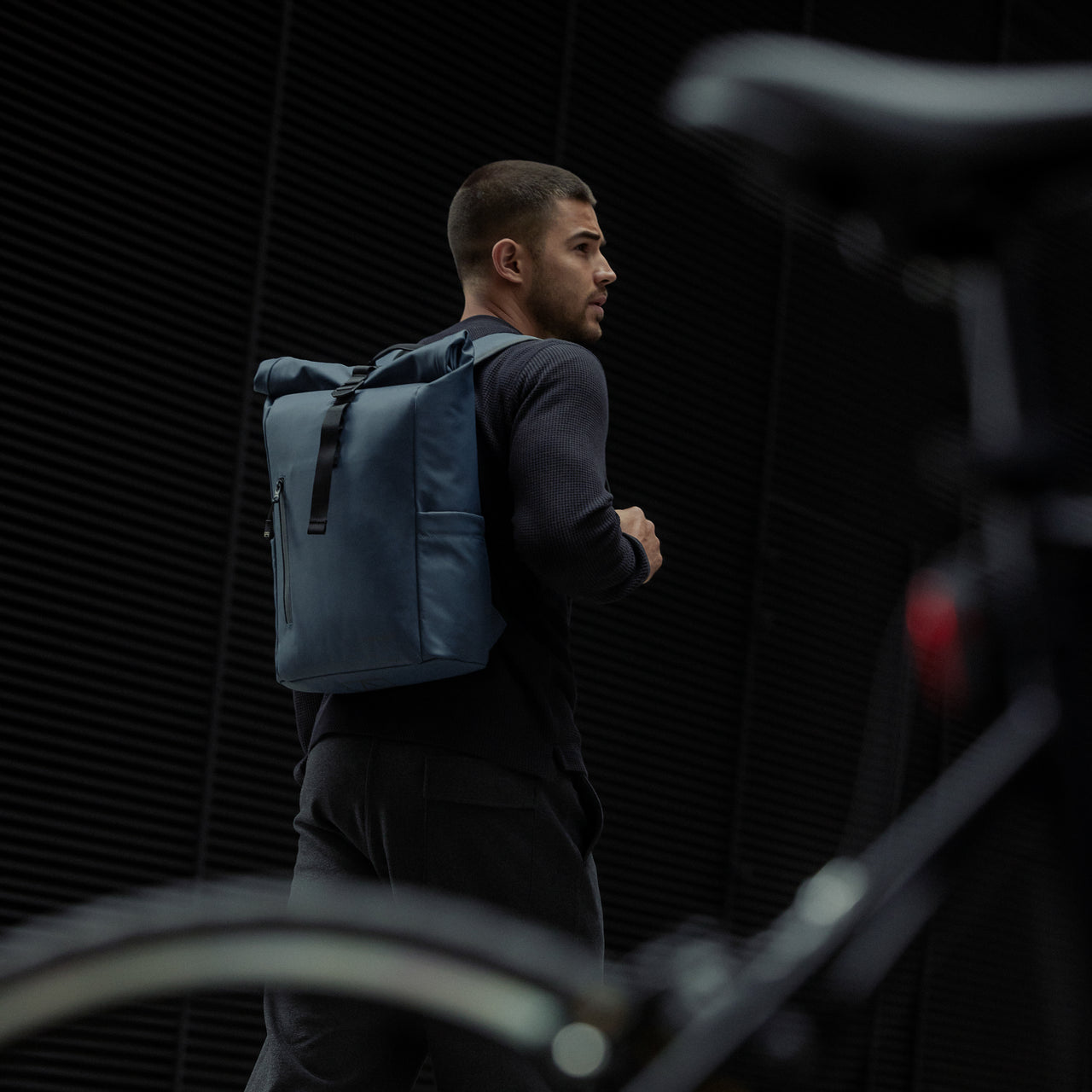 A man standing next to a bike with a Roll Top 15L backpack in Tasmin Blue on his back