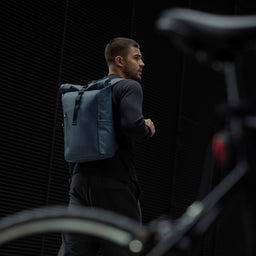 A man standing next to a bike with a Roll Top 15L backpack in Tasmin Blue on his back