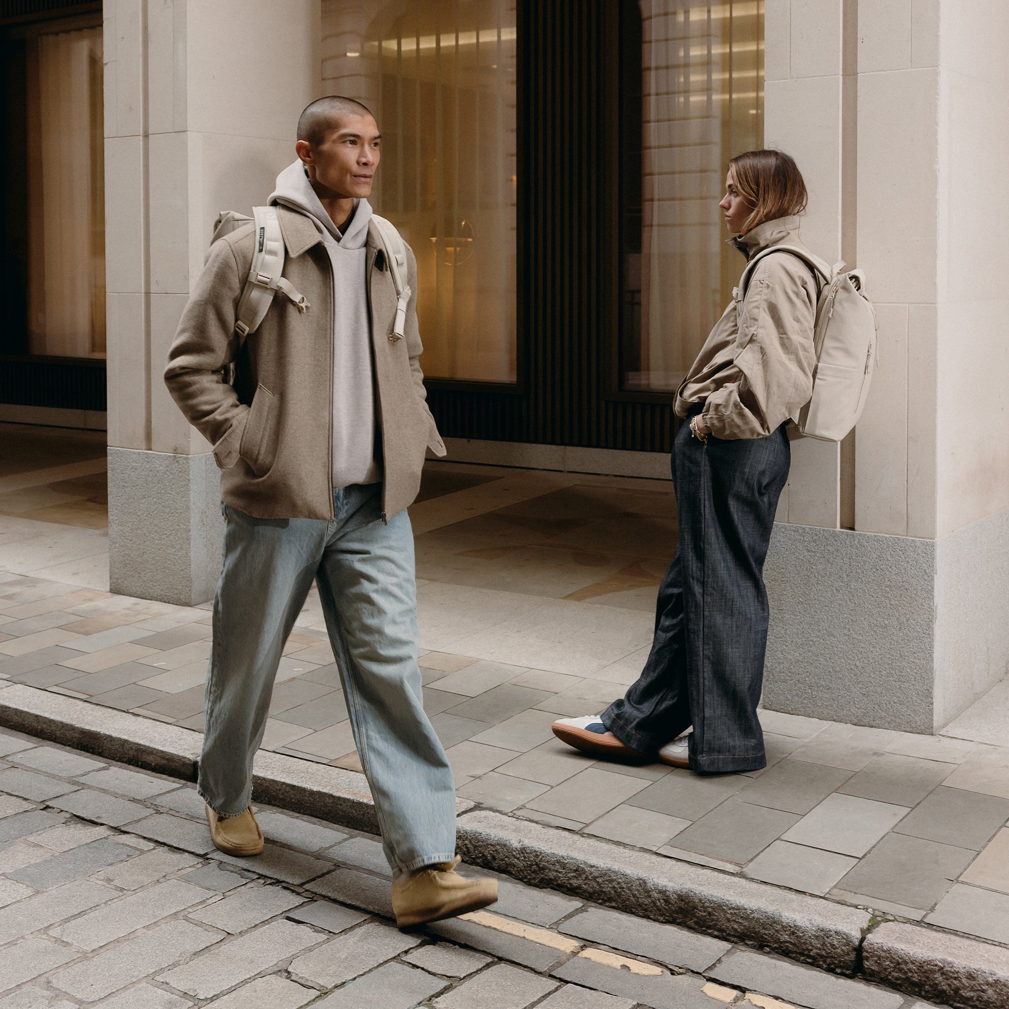 A man and woman standing on a pavement wearing Roll Top backpacks on their backs in Tonal Sand