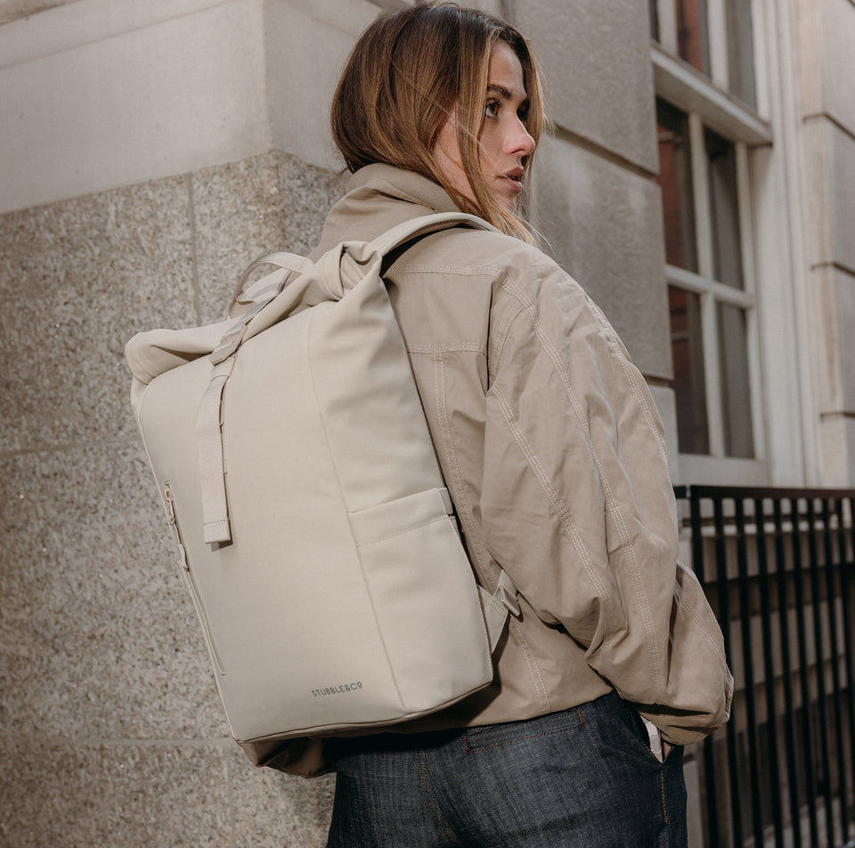 A woman standing on the pavement in front of a brick wall wearing a roll top15l in tonal sand on her back