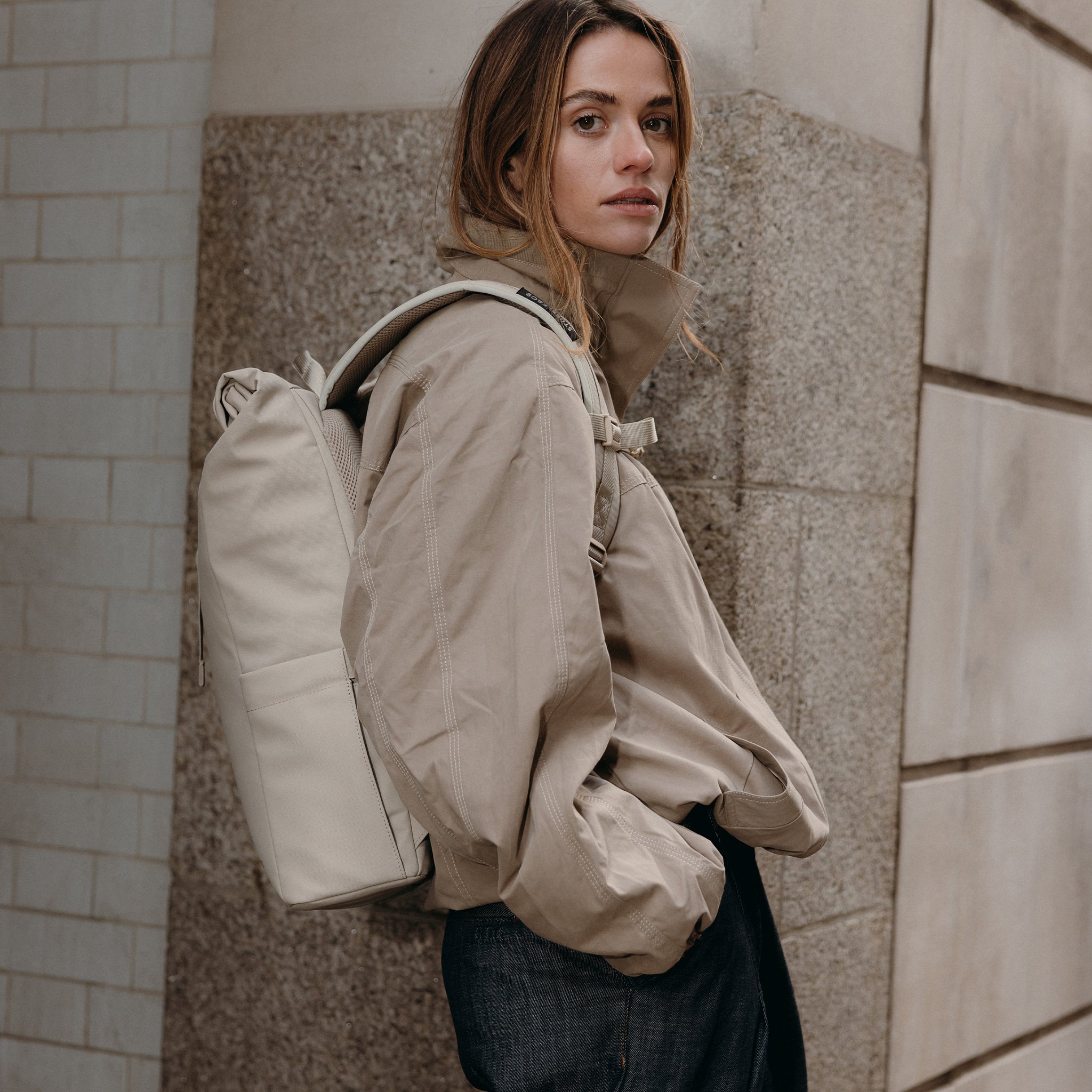A woman leaning against a wall wearing a Roll Top 15l backpacks on her back in Tonal Sand