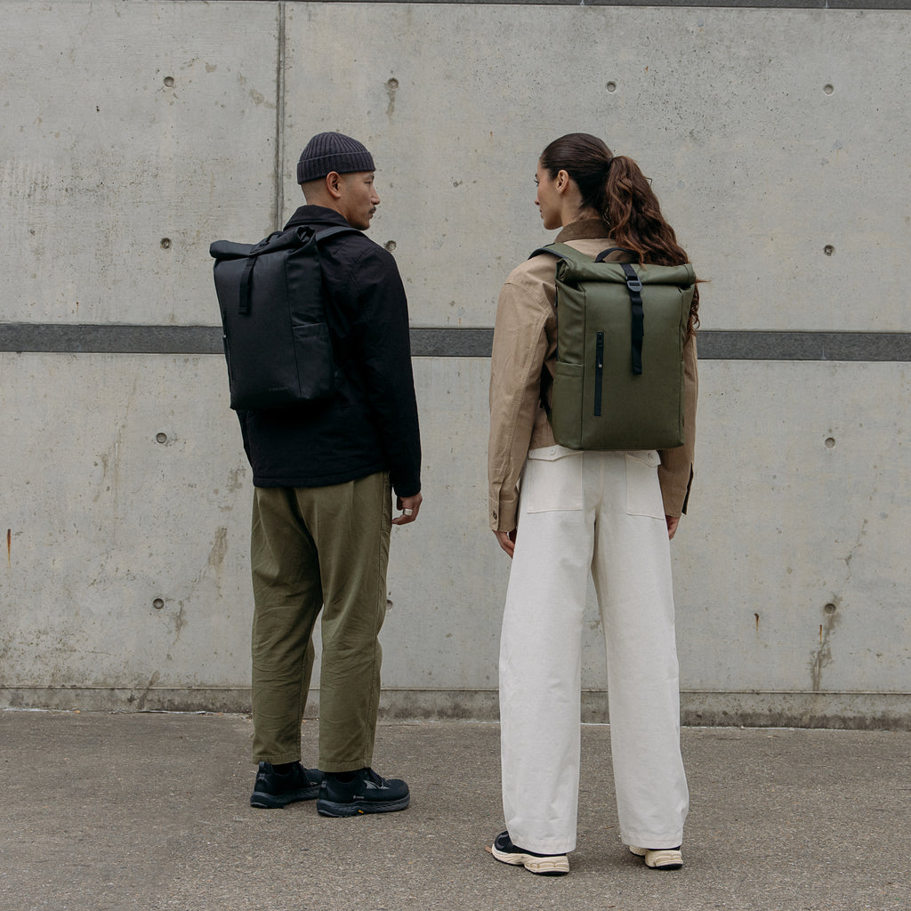 A man and woman standing next to each other wearing Roll Top 15L backpacks on their backs in black and Urban Green