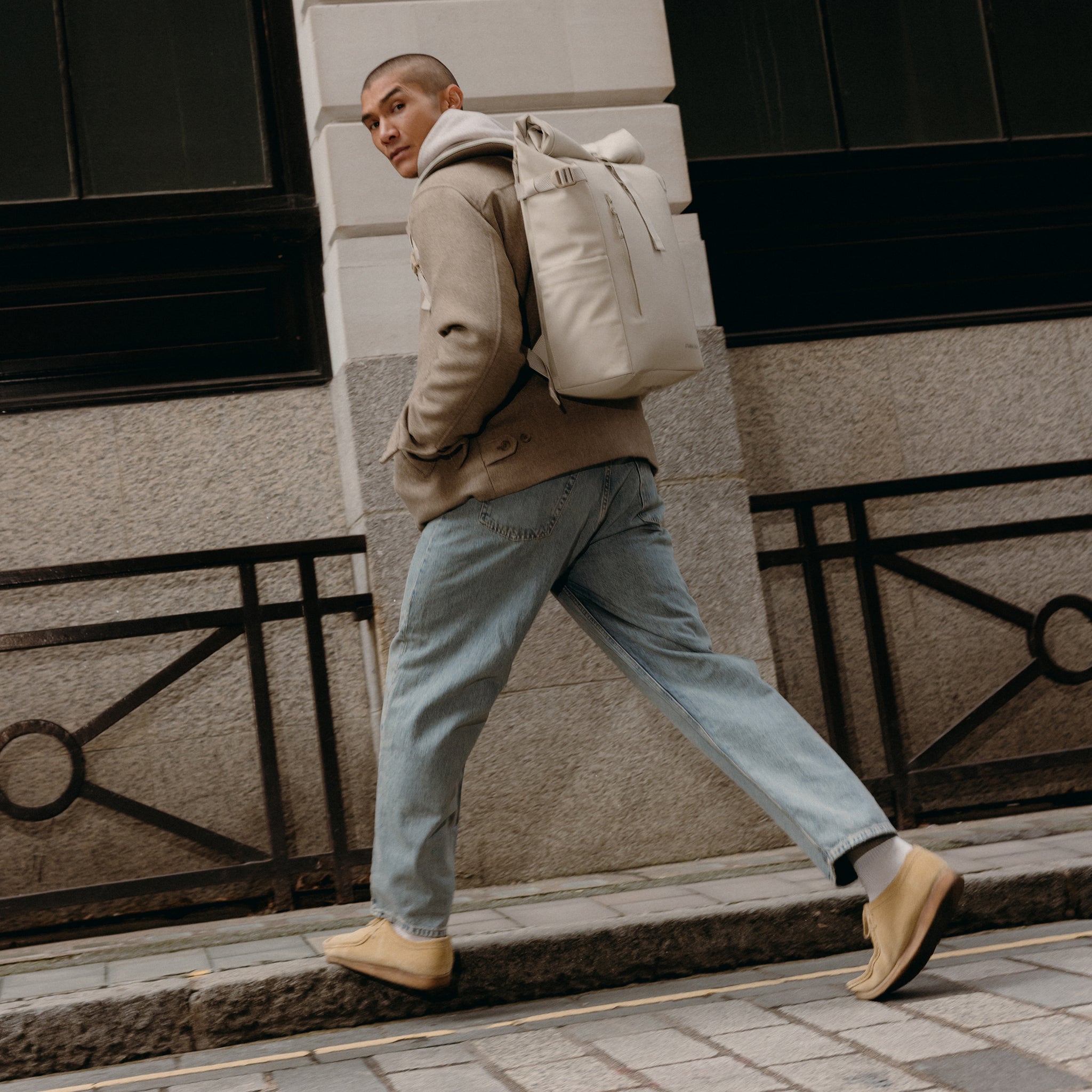 A man walking on the pavement looking at the camera wearing a Roll Top 20L backpack in Tonal Sand on his back