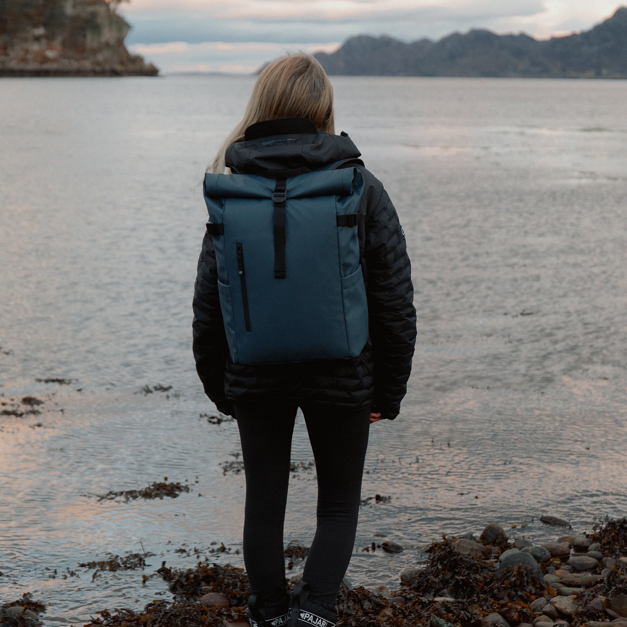 Women wearing The Roll Top 20L backpack in Tasmin Blue by water