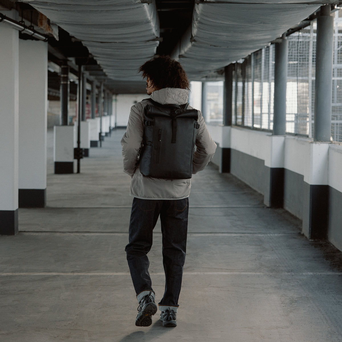 Back view of a man walking down a covered walkway wearing The Roll Top 20L in All Black