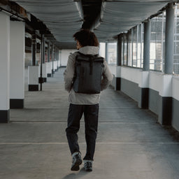 Back view of a man walking down a covered walkway wearing The Roll Top 20L in All Black