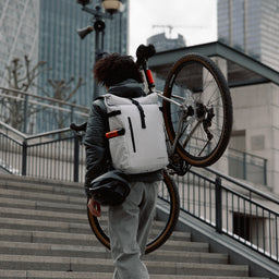 Man wearing The Roll Top 20L in Arctic White carrying a bike up steps
