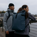 A man and woman looking over a river wearing Tasmin Blue Roll Top 20L backpacks on their backs