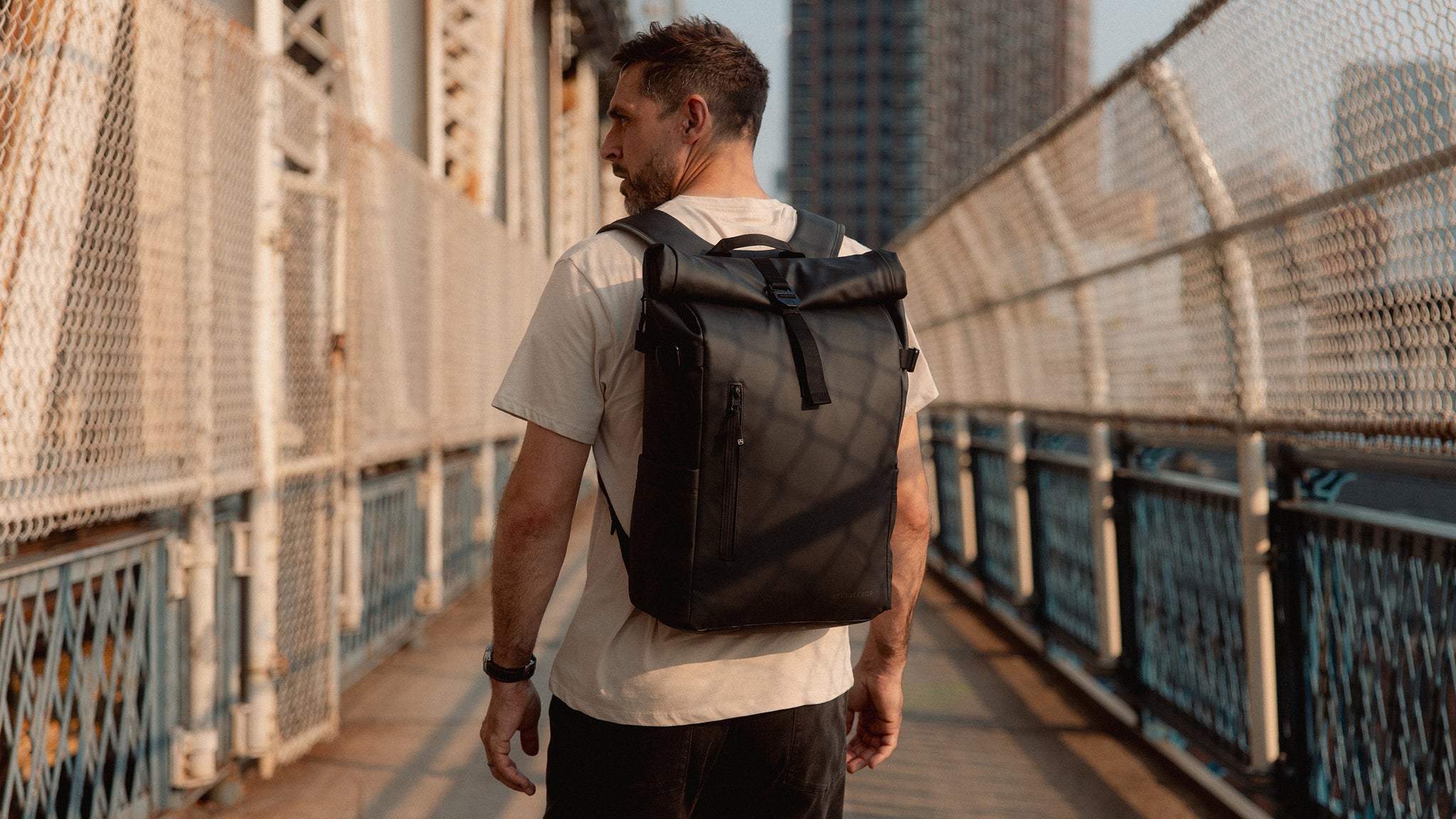 A man wearing a Black Roll Top 20L as he is crossing a bridge