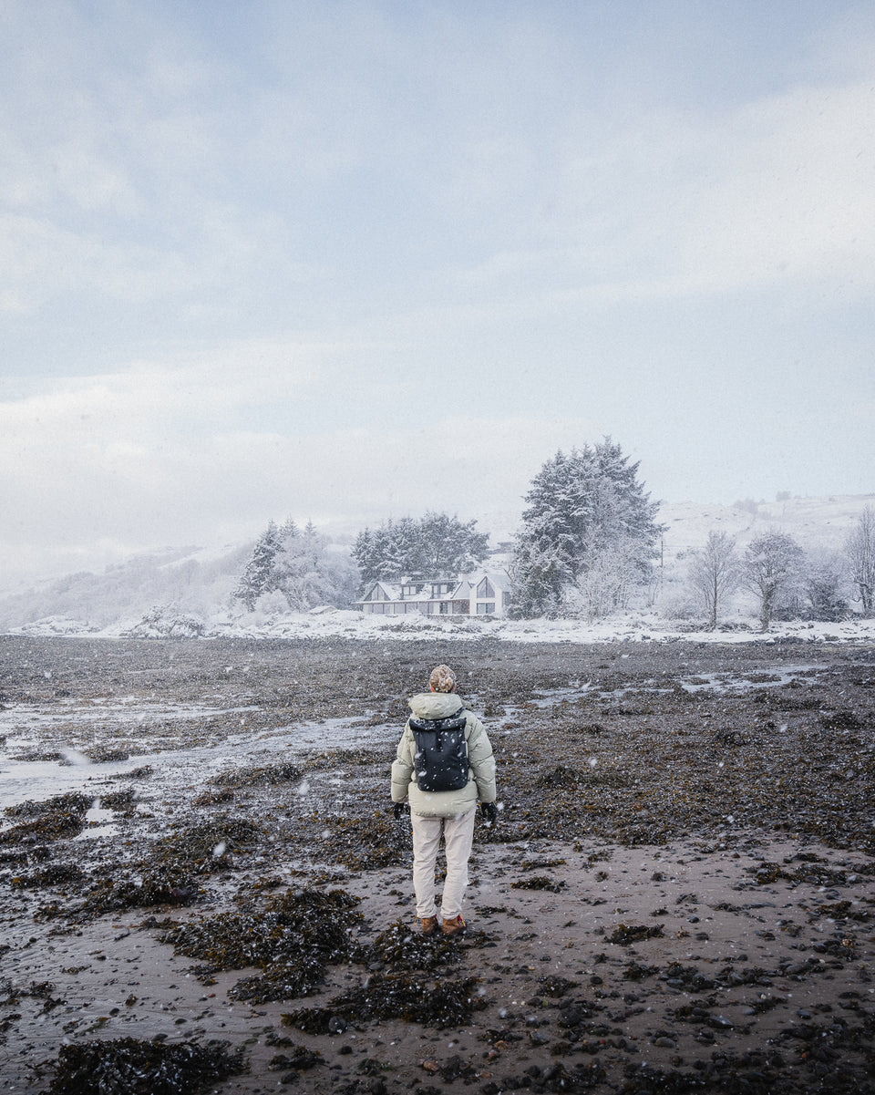 A woman standing in the snowy countryside wearing a roll top 20l in tasmin blue on her back