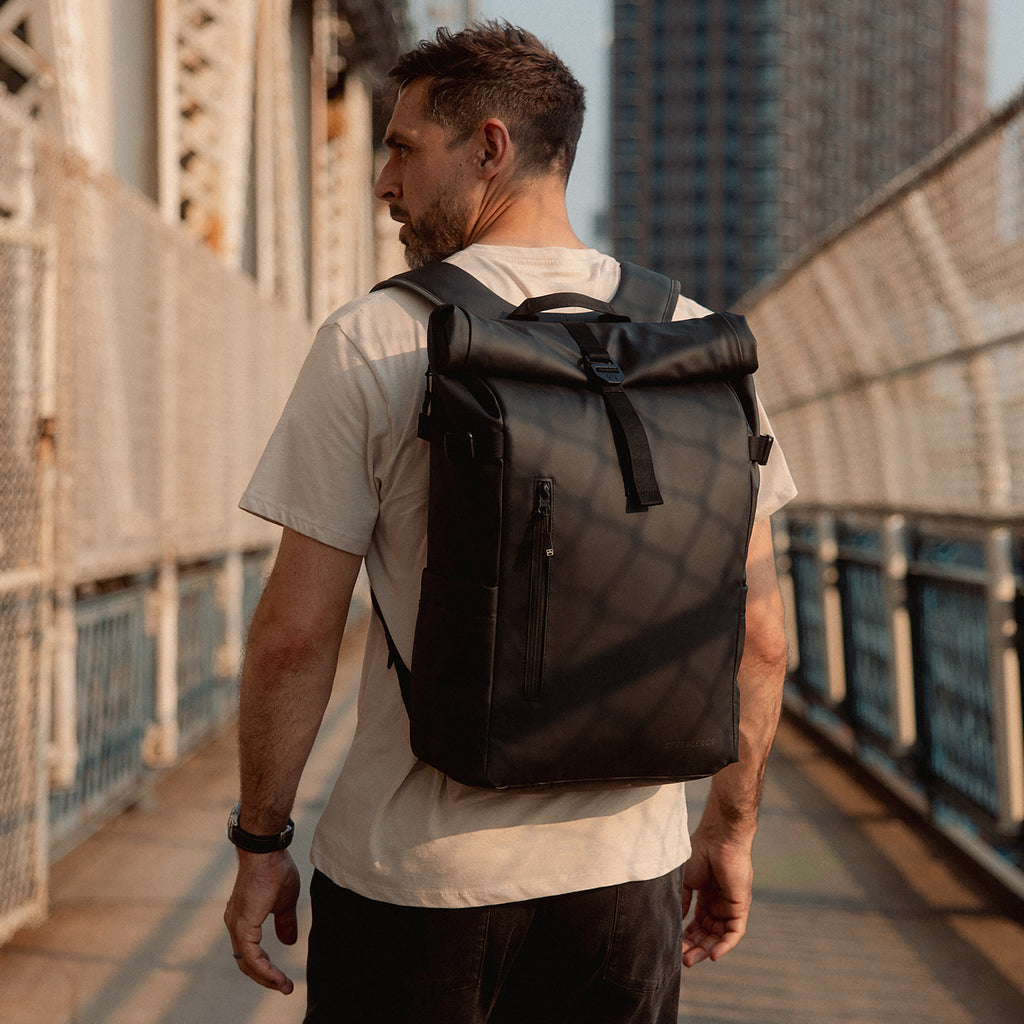 A man walking across a bridge looking to his side whilst wearing a Roll Top 20L in All Black on his back
