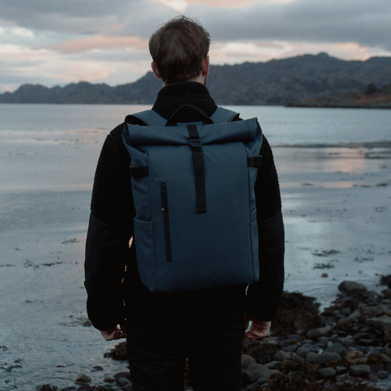 Man wearing The Roll Top 20L backpack in Tasmin Blue by the sea