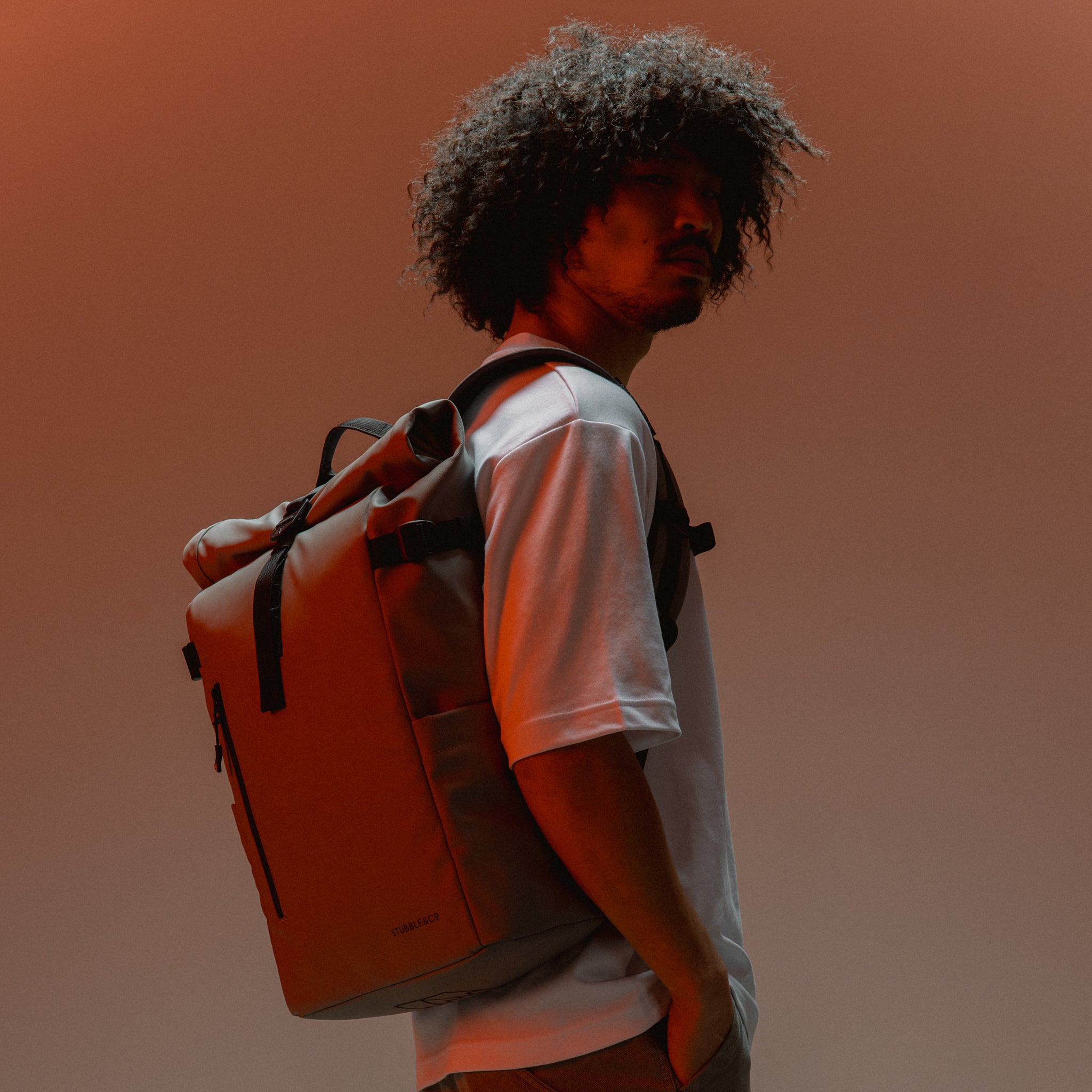 A man wearing a concrete roll top backpack on his back in a studio with an orange light 