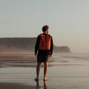 Man wearing The Roll Top 20L backpack in Ember Orange on a beach