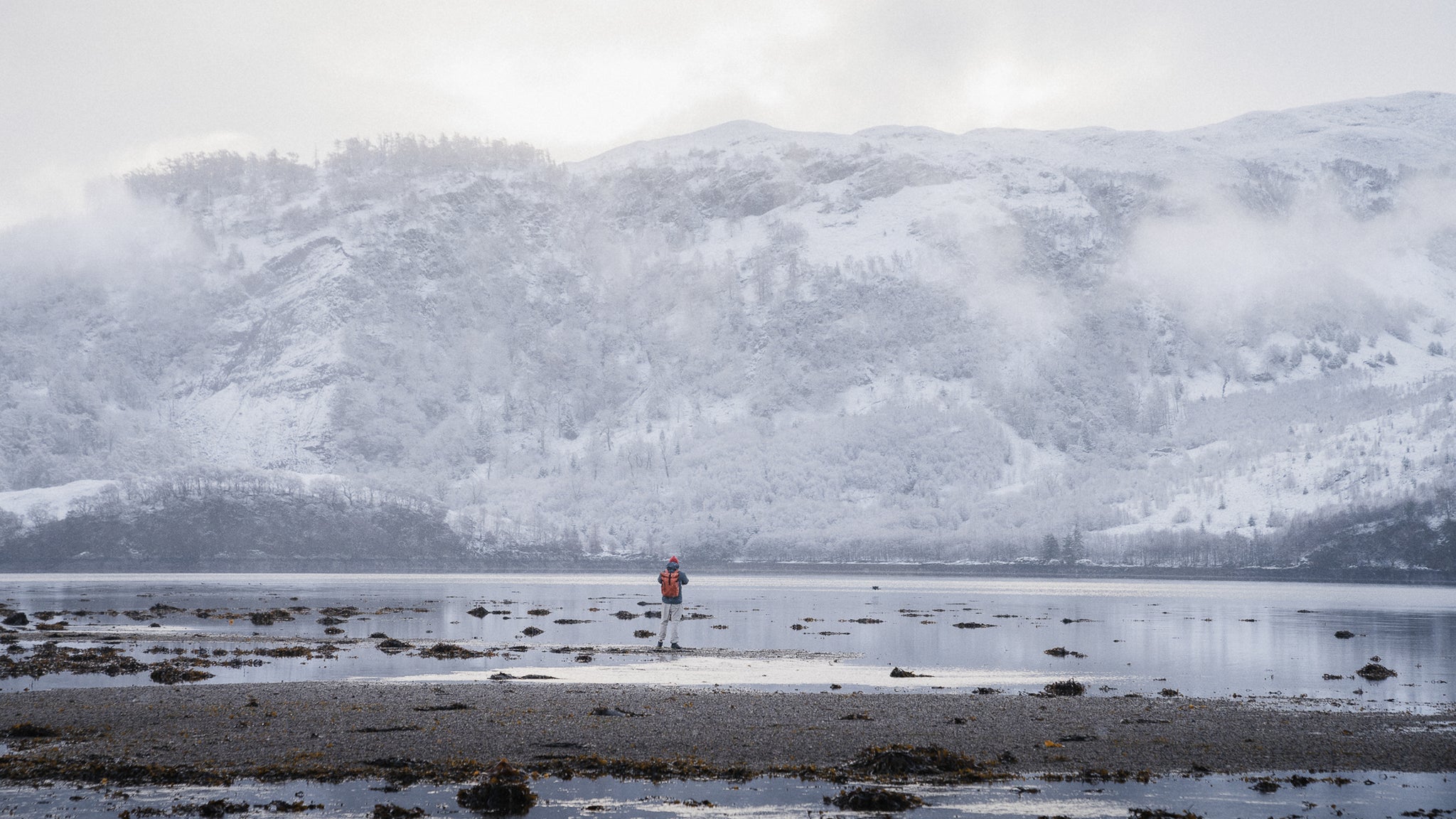 a man standing by a snowy lake and landscape wearing a roll top 20l on his back in rust