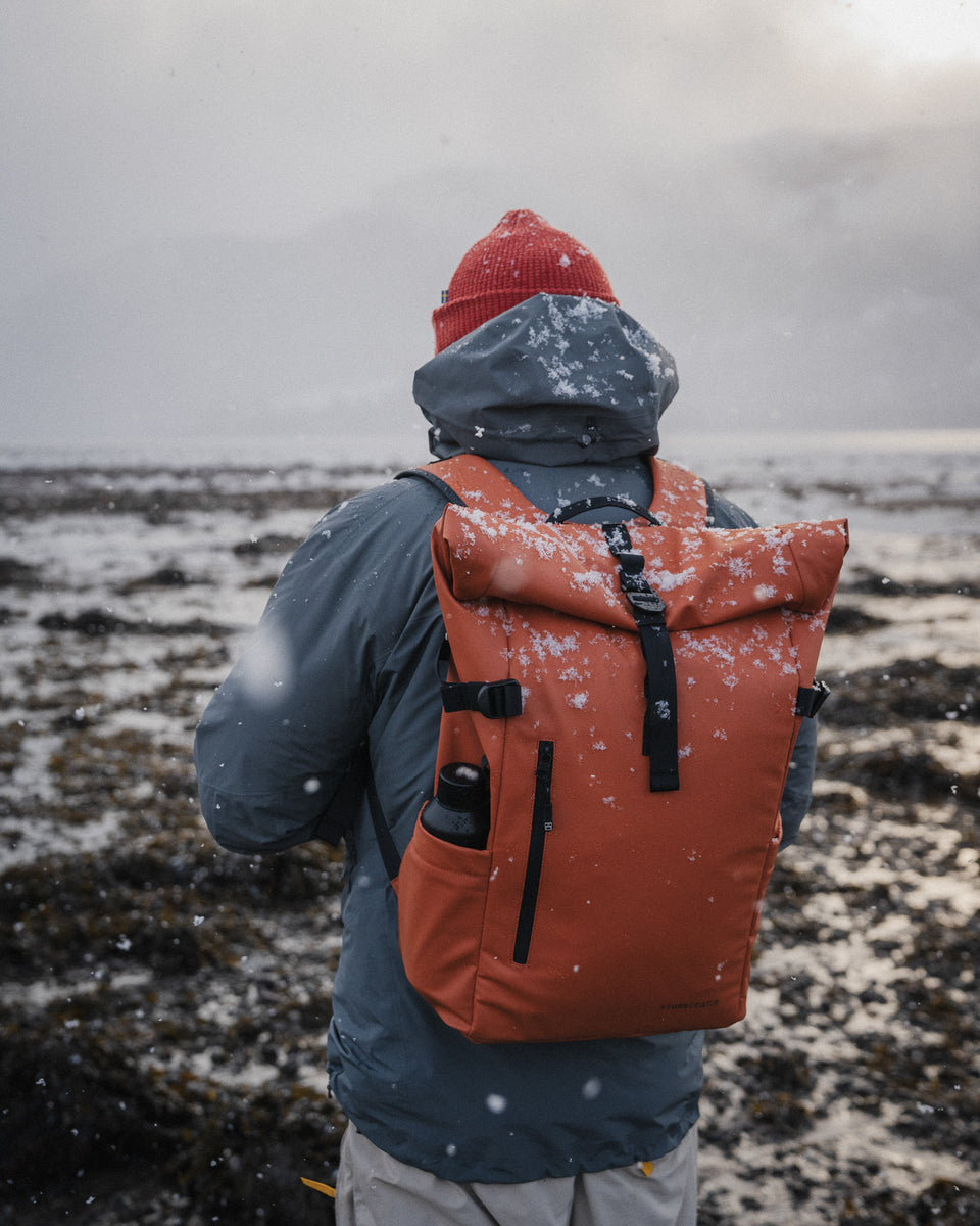 A man standing in the snow with a roll top 15l in rust on his back