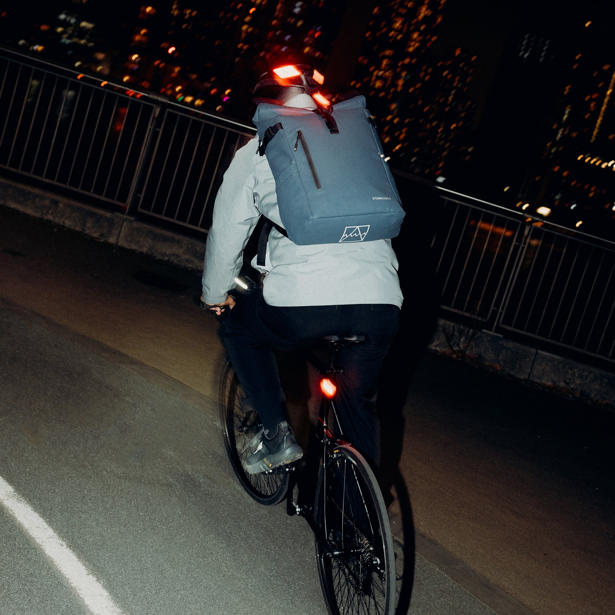 A man cycling at night wearing a Roll Top 20L in Tasmin Blue backpack on his back with a bike light attached