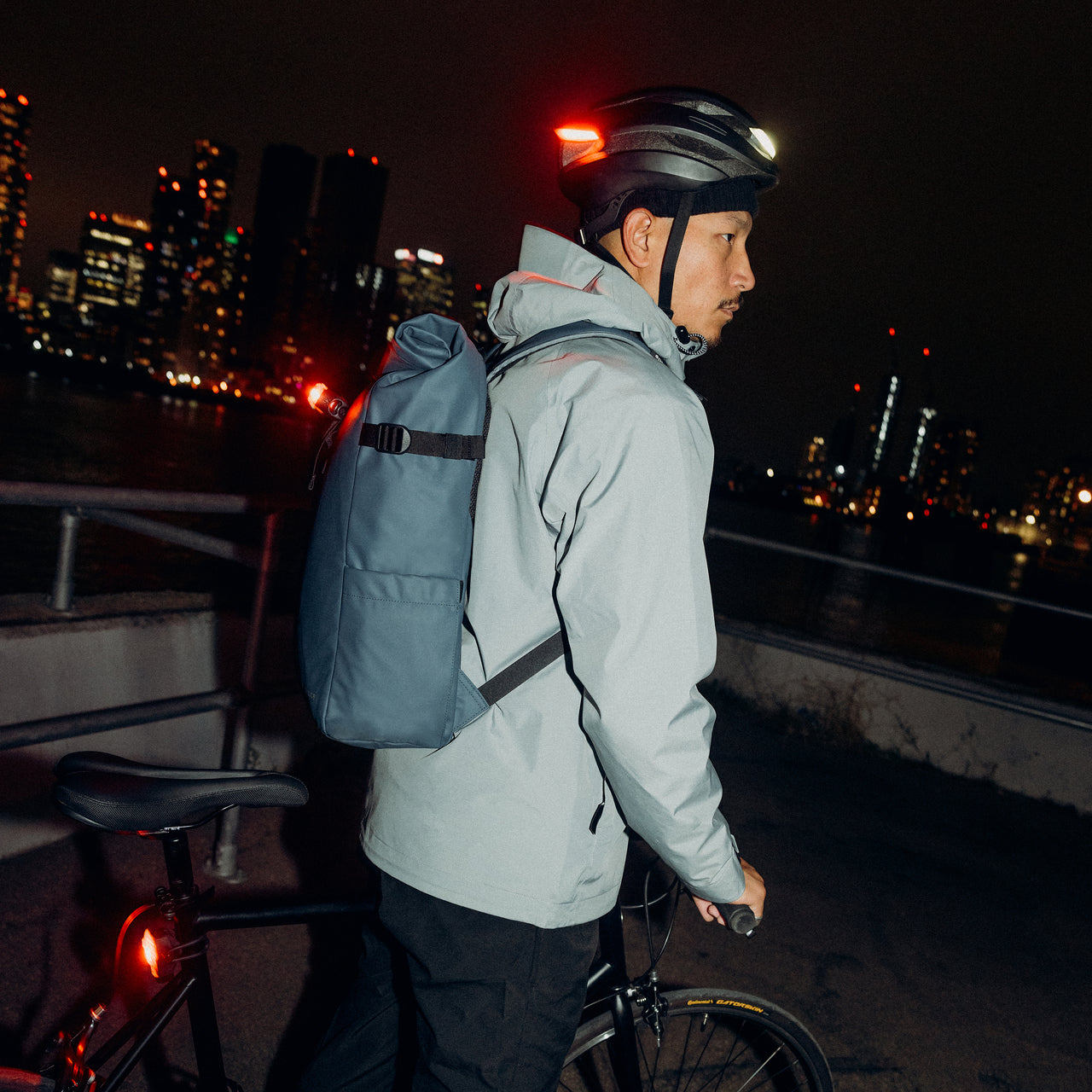 A man standing with his bike wearing a Tasmin Blue Roll Top 20L on his back with bike lights on