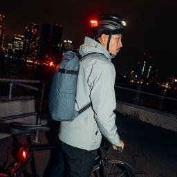 A man standing with his bike wearing a Tasmin Blue Roll Top 20L on his back with bike lights on