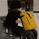 a man sitting in a tube station whilst placing a laptop into the laptop compartment into a Yellow roll top 20l