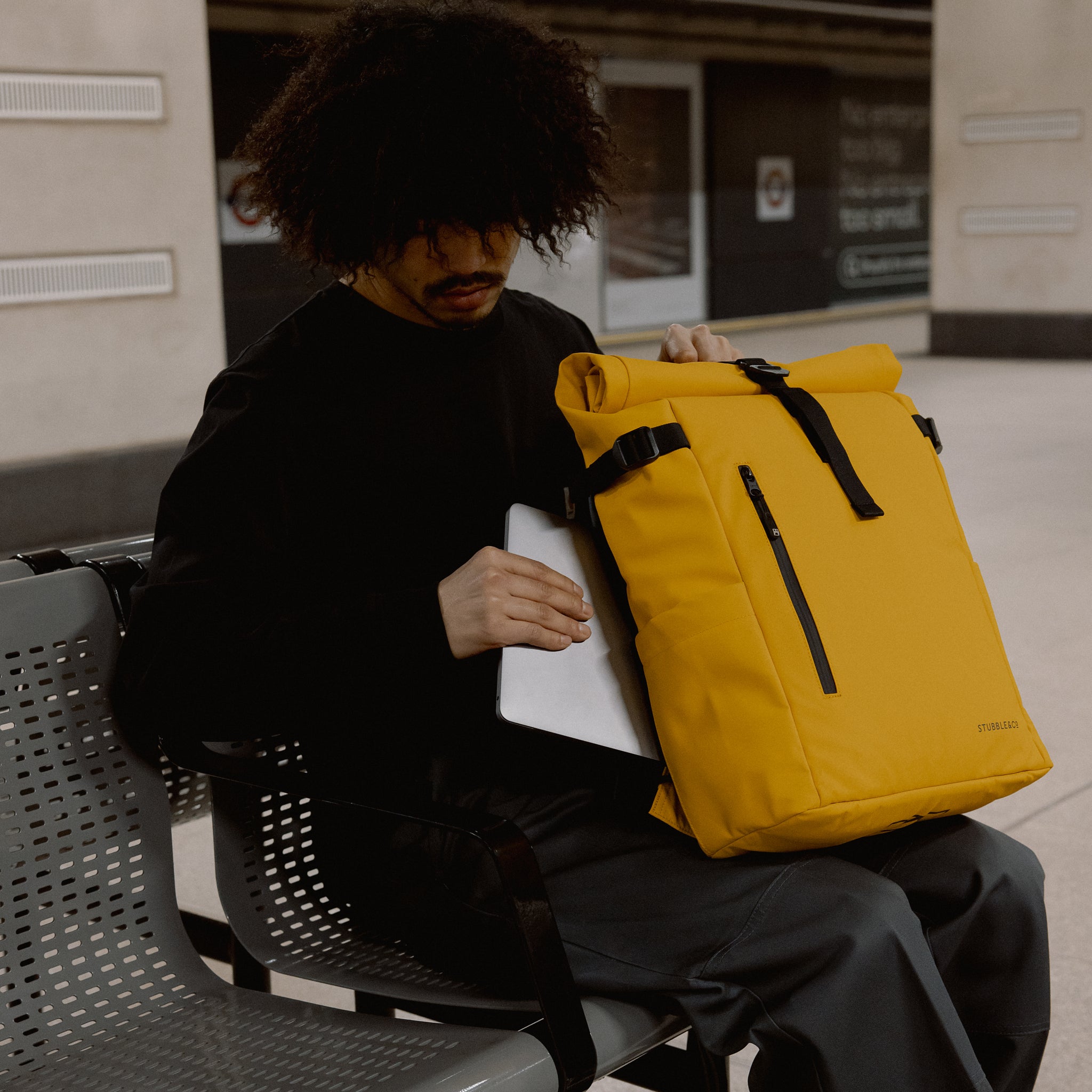 a man sitting in a tube station whilst placing a laptop into the laptop compartment into a Yellow roll top 20l