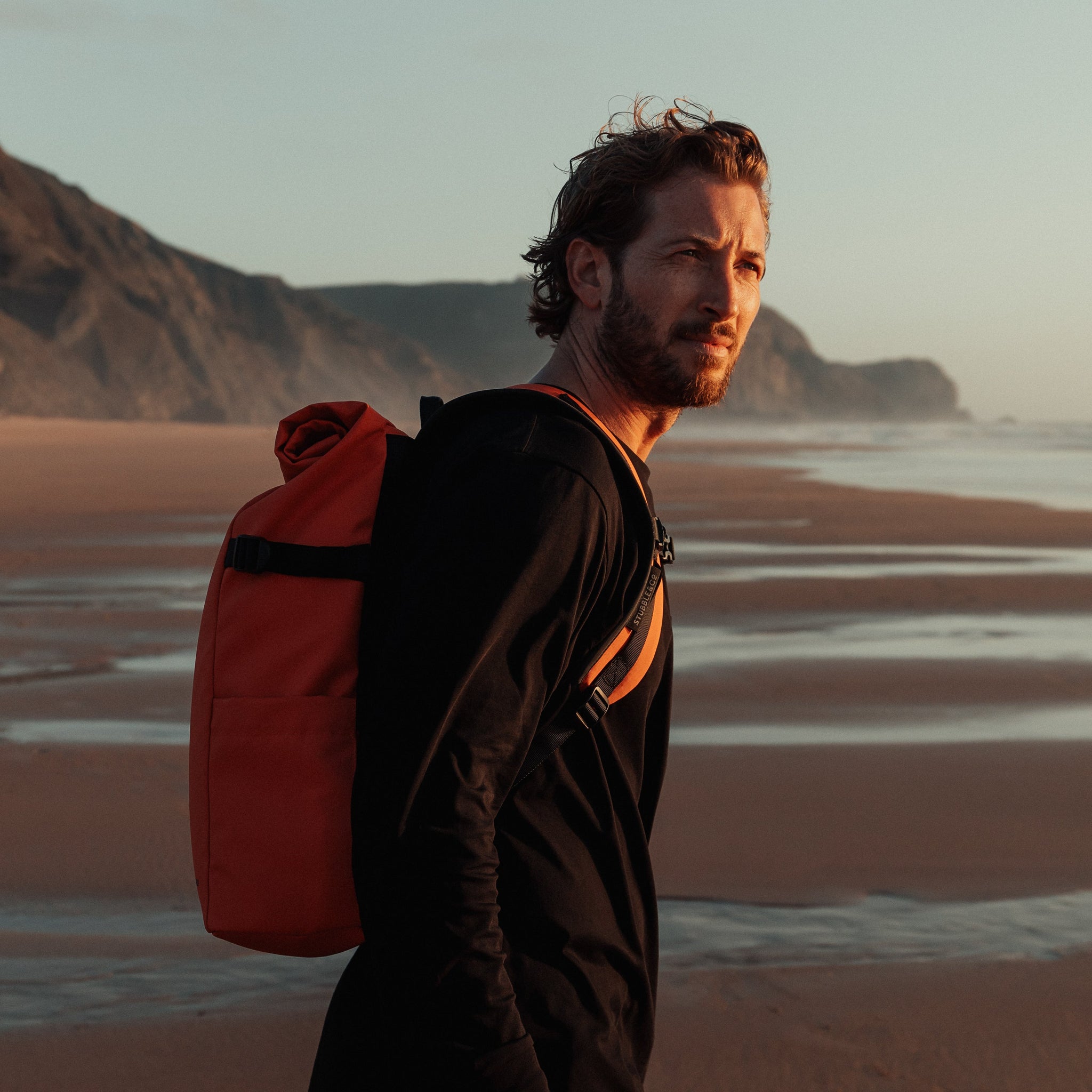 Man wearing The Roll Top 20L backpack in Ember Orange on a beach