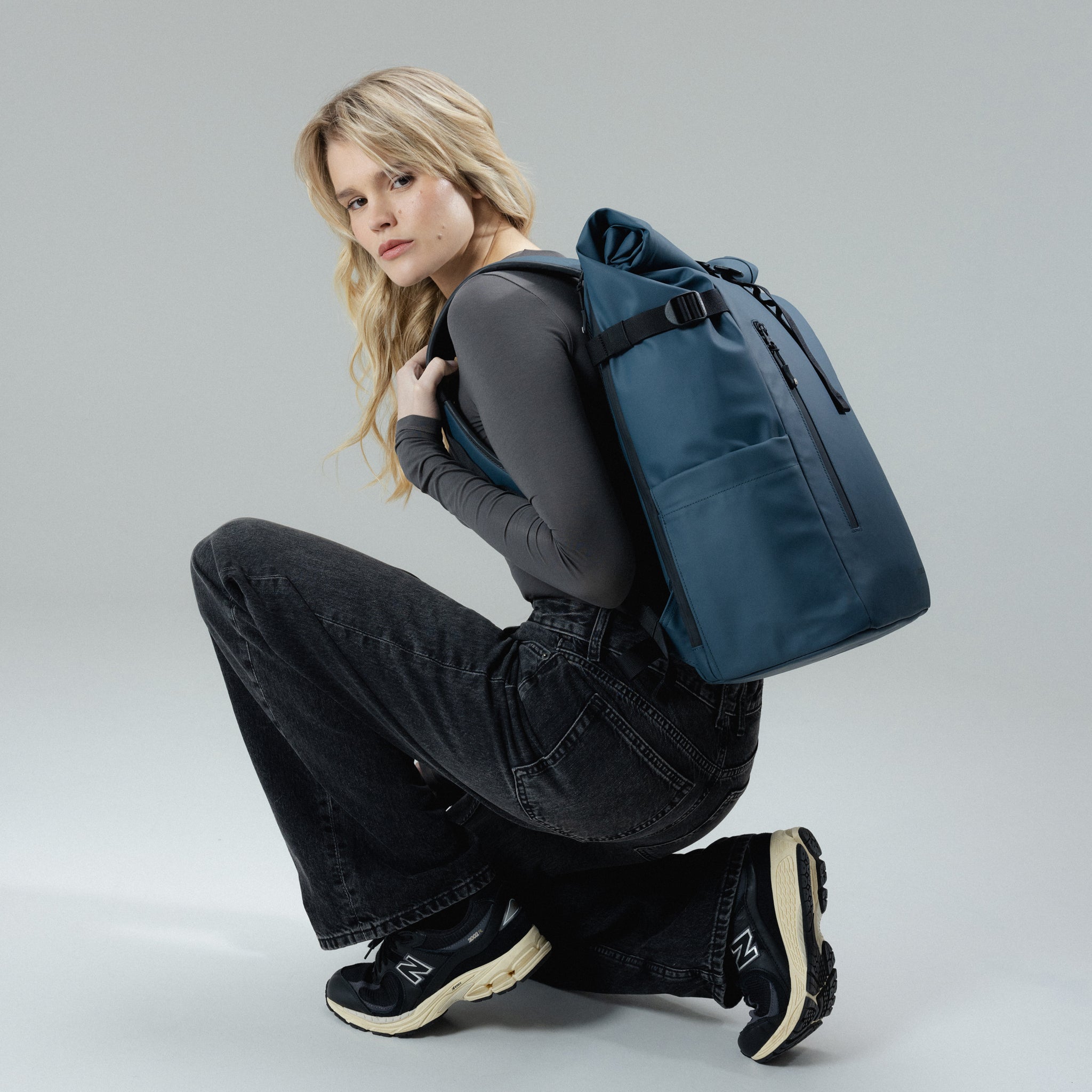 A woman crouching down wearing a Roll Top 30L on her back in Tasmin Blue