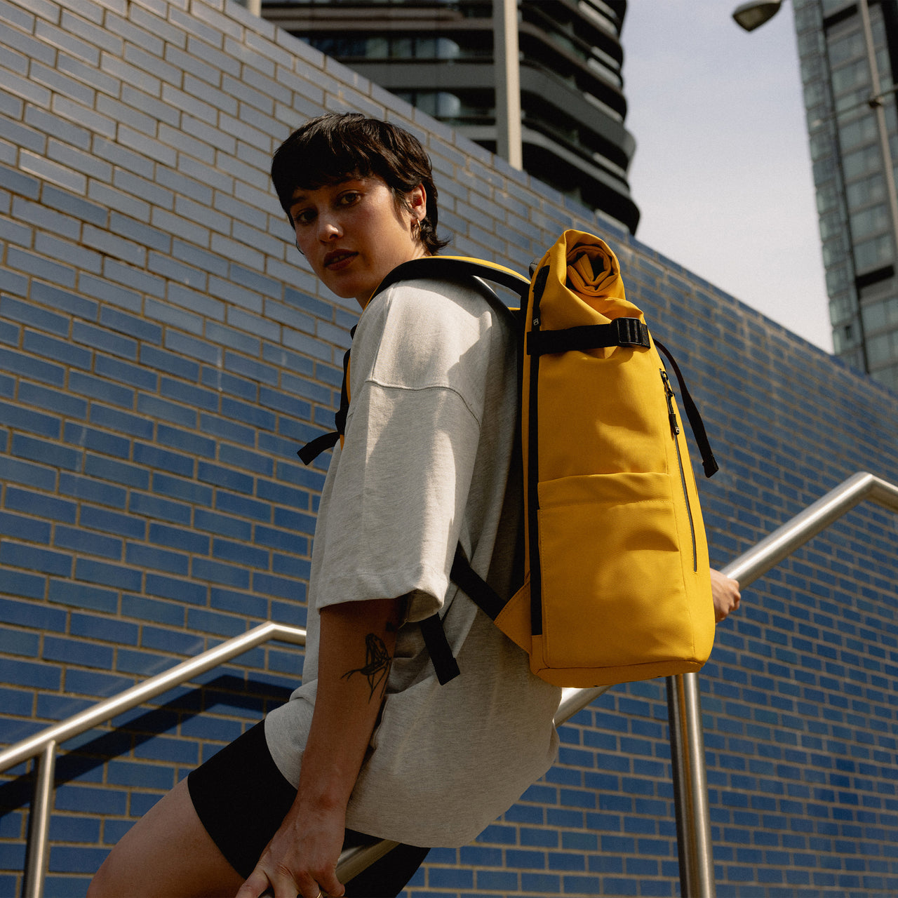 A woman sitting on the bannister of stairs wearing a Roll Top 30L in Yellow on her back
