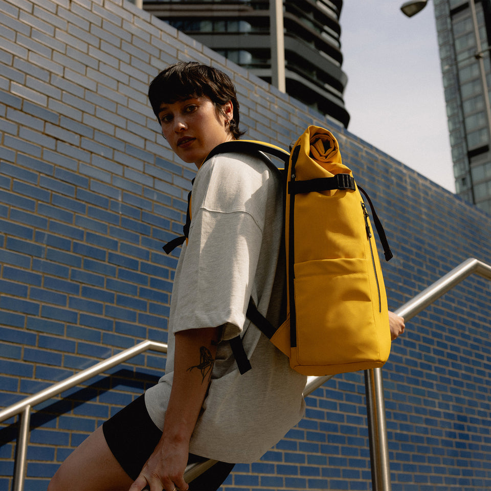 A woman sitting on the bannister of stairs wearing a Roll Top 30L in Yellow on her back