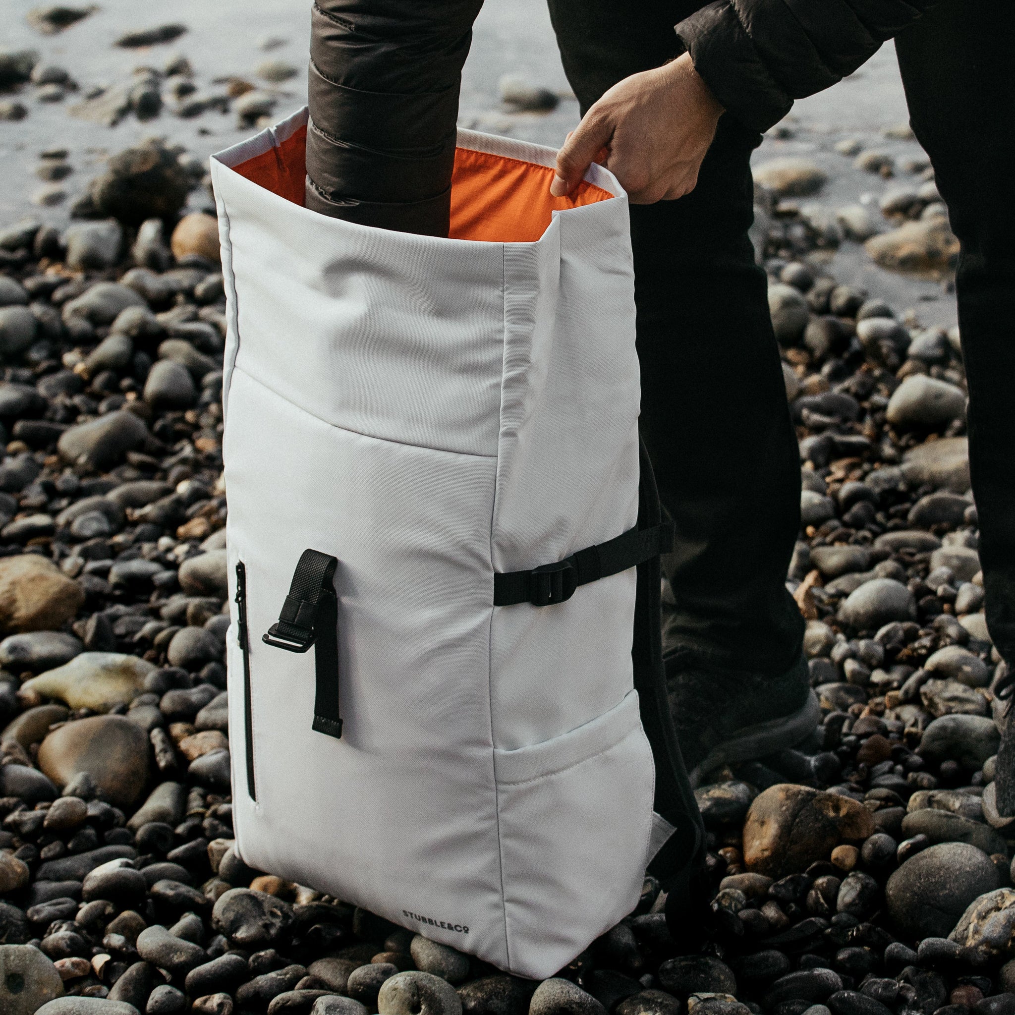 The Roll Top in Arctic White on a pebble beach with someone reaching inside the open bag.
