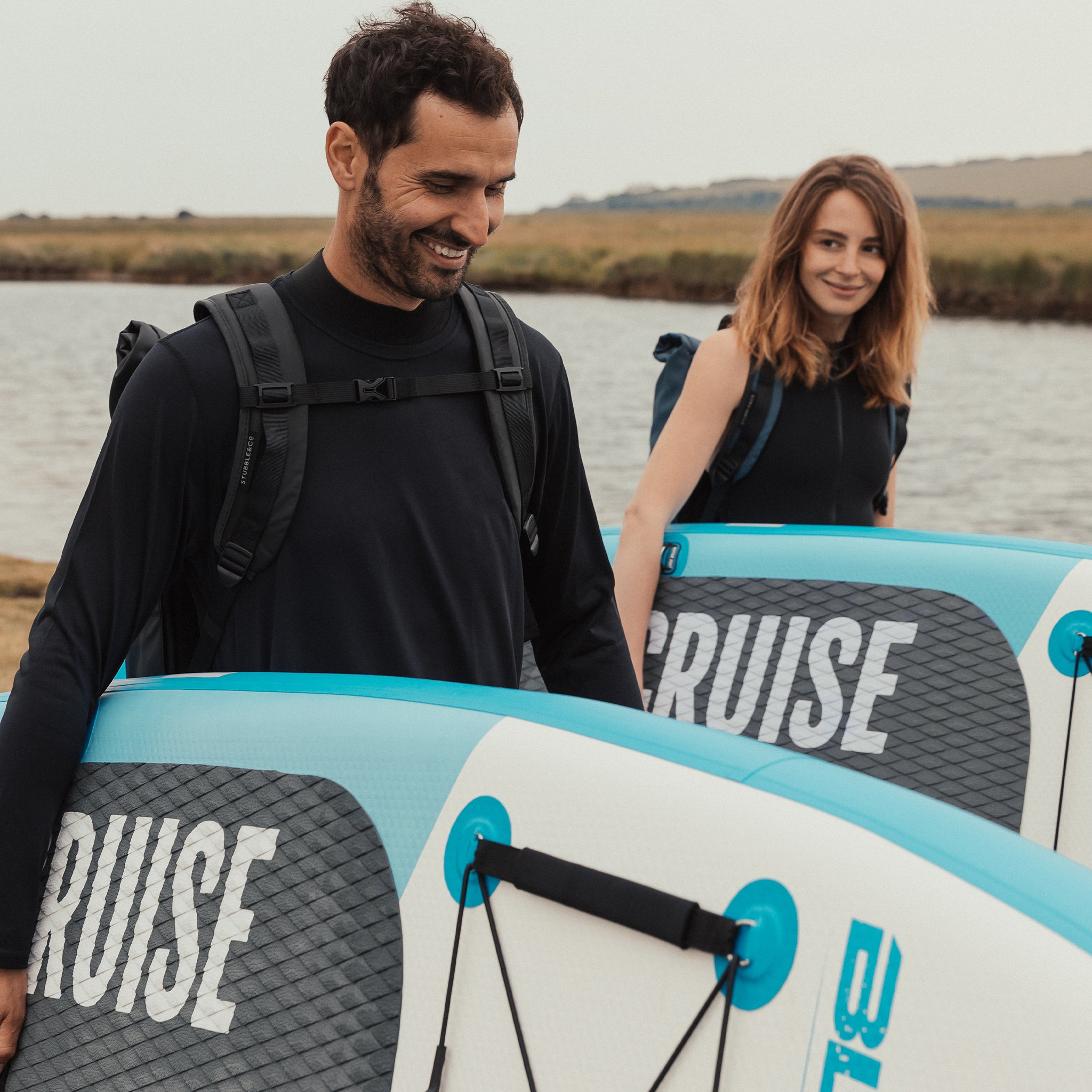 A couple carrying stand up paddle boards wearing roll tops on their backs