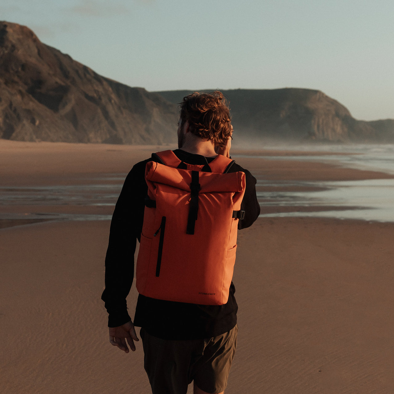 Man walking away from camera on the beach wearing The Roll Top in Ember Orange