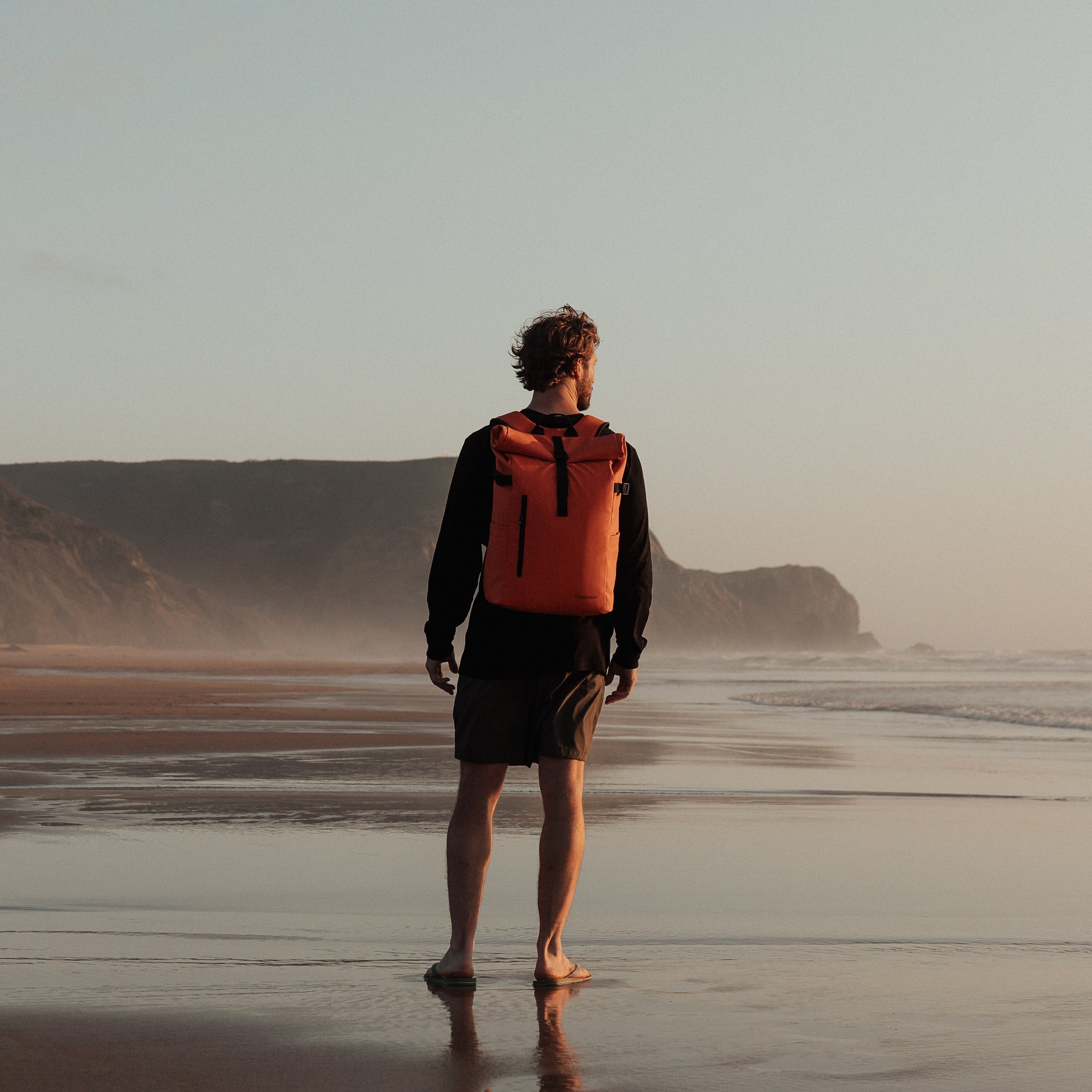 Man standing on beach with back to camera wearing The Roll Top in Ember Orange